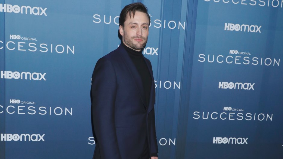 Kieran Culkin poses in front of a blue Succession backdrop while wearing a navy suit.