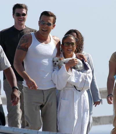 Ben Affleck wearing a white tank top and khakis next to Jennifer Lopez in a white robe while holding a dog in 2002.