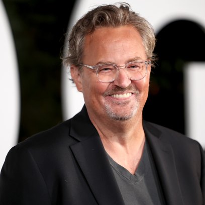 Matthew Perry smiles while wearing a gray tshirt and black jacket