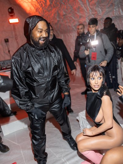 Bianca Censori sits while wearing a black bodysuit with nothing underneath next to Kanye West in an all black ensemble.