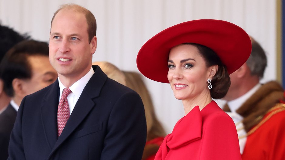 Prince William wearing a dark suit and red tie next to Kate Middleton wearing a red ensemble with a matching hat.
